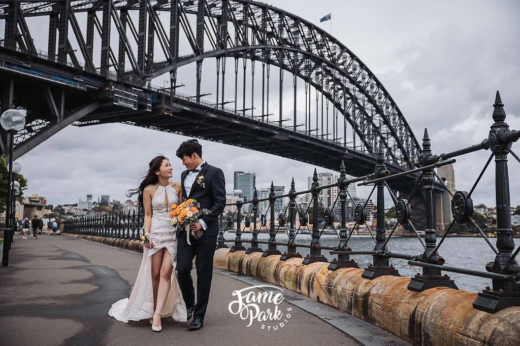 The bride and groom are walking side by side under the Sydney Harbour Bridge