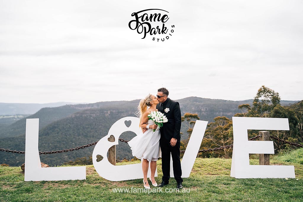 Against the stunning backdrop of the Blue Mountains, the bride and groom share a tender kiss in front of a beautifully adorned "Love" sign, symbolizing their love amidst the breathtaking natural scenery.