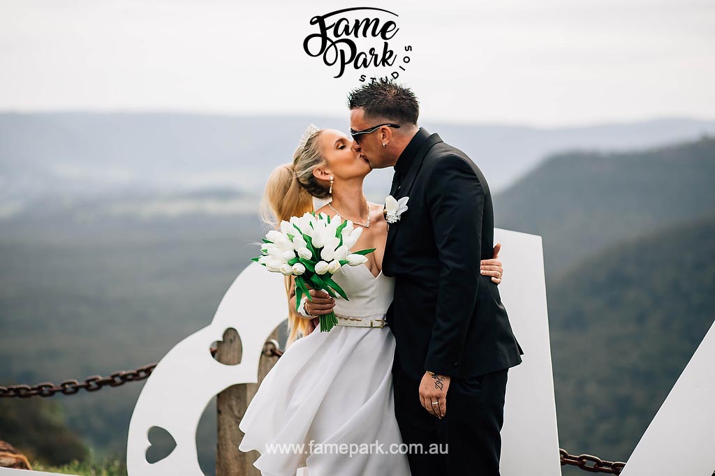 In a picturesque setting surrounded by the majestic Blue Mountains, the bride and groom embrace in a passionate kiss, framed by a charming "Love" sign, creating a moment of romance and beauty that blends nature and love harmoniously.