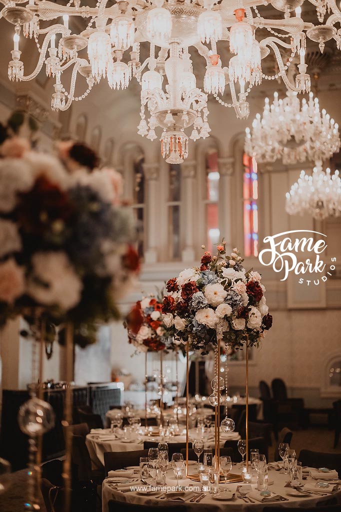 The reception design is characterized by a neutral color palette, refined table settings, and the gentle glow of elegant chandeliers, casting a warm and inviting light upon the lifelike silk flower arrangements