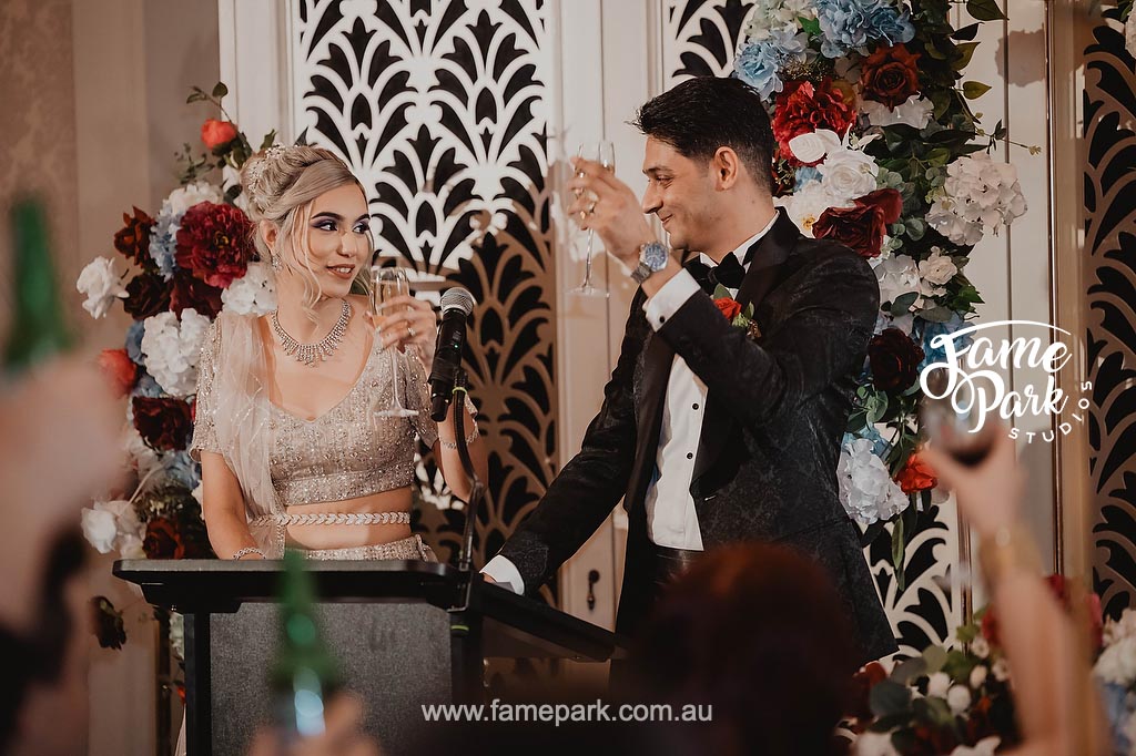 The newlyweds raise their glasses high in a celebratory toast, their faces beaming with happiness and love as they share a heartfelt moment with their guests.