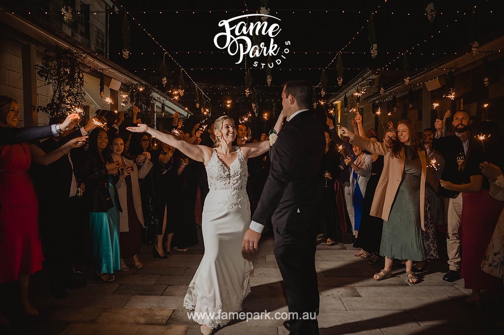 The bride and her husband gracefully sway on the dance floor, surrounded by their guests holding sparklers, creating a magical ambiance of twinkling lights and celebratory energy.