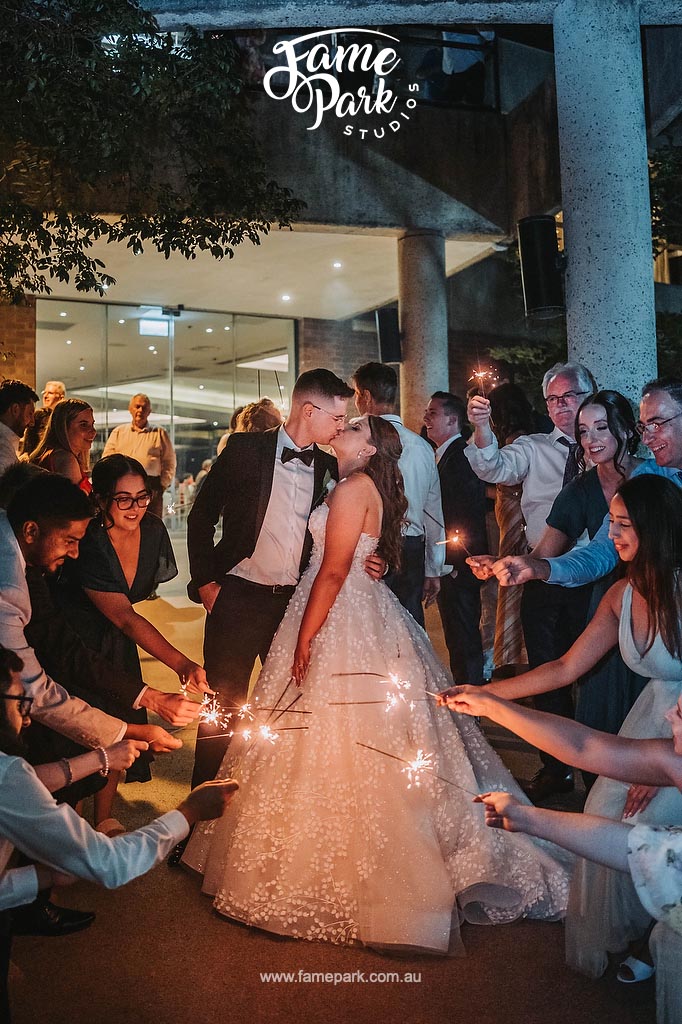 Amidst the radiant glow of sparklers, the bride and groom steal a passionate kiss, their guests forming a sparkling backdrop, symbolizing the shared excitement and well wishes surrounding their union.
