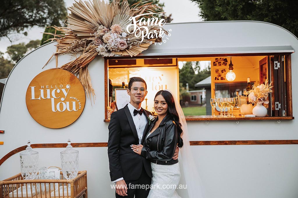 A bride and groom standing in front of a vintage trailer at their wedding reception cocktail party.