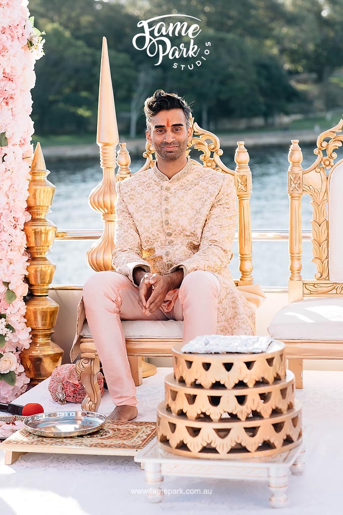 An Indian man dressed in traditional wedding attire sits on a gold throne.